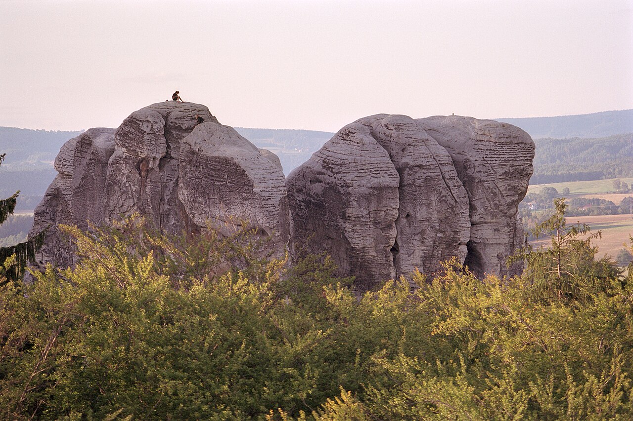 Two large sandstone rock towers above green forest in the Bohemian Paradise, with fields and low hills behind