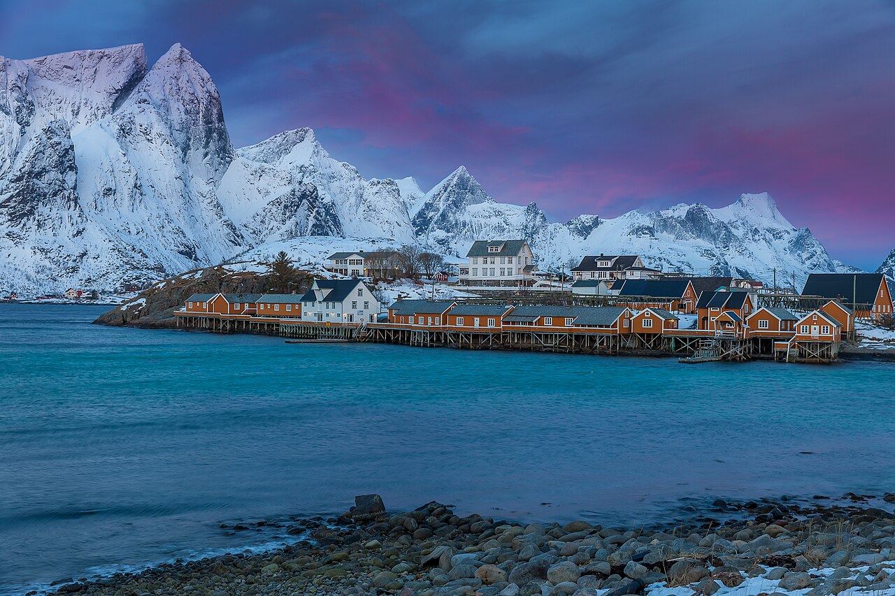 Red fishing cabins on stilts by the water below steep mountains in the village of Sakrisøy in the Lofoten Islands