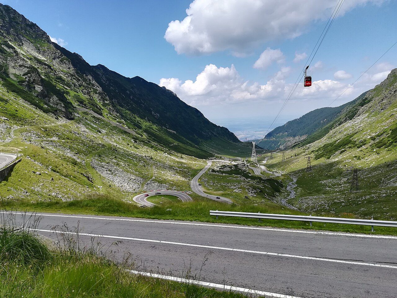 A road winding in hairpin bends down a green Carpathian valley on the Transfăgărășan, with a cable car overhead