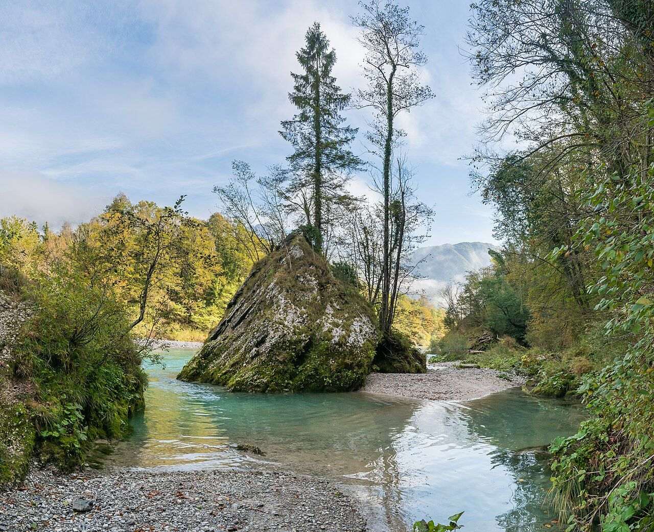 A turquoise stretch of the Soča river with a large mossy rock, a gravel bank and forested hills in Slovenia