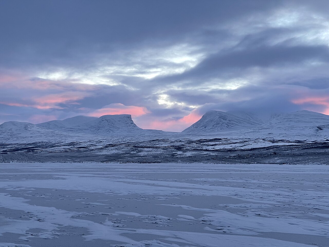 Snow-covered mountains under a pink and gray winter sky above a frozen lake at Abisko in Swedish Lapland
