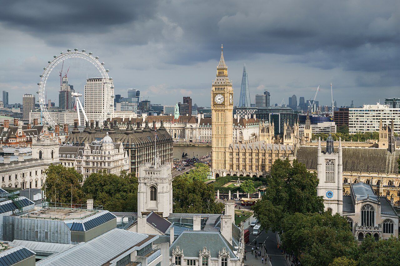The Palace of Westminster and the Elizabeth Tower, known as Big Ben, seen from above across the rooftops, with the London Eye on the skyline under a gray sky