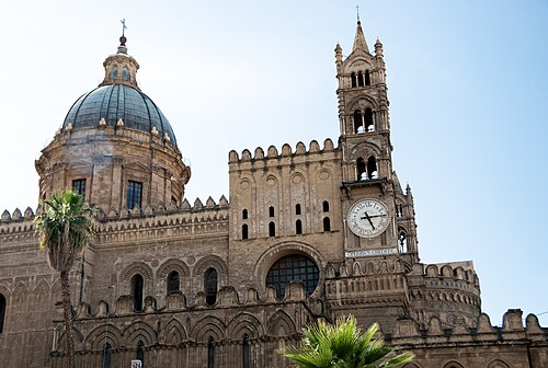 Palermo Cathedral, its dome and clock tower against a blue sky, with palm trees in front