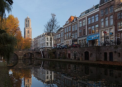 The Dom Tower rising above brick canal houses and an arched bridge on the Oudegracht in Utrecht, with autumn trees and reflections in the water